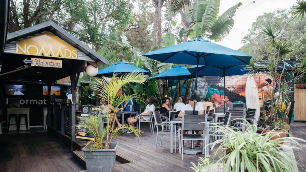 5 Best Hostels in Noosa 5 Travellers sit on an outdoor deck with blue umbrellas at Nomads, one of the best hostels in Noosa