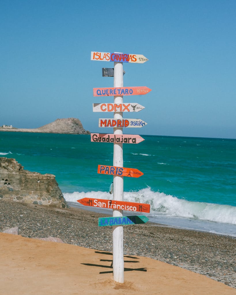 The Quintessential Baja California Sur Road Trip Itinerary 16 Colorful wooden signpost on a rocky beach in Cabo Pulmo points to cities like Paris, San Francisco, and CDMX with ocean waves in the background.