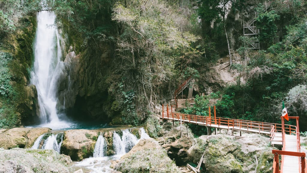 How to Visit Cascada El Aguacate: San Luis Potosi, Mexico 9 Cascada El Aguacate in Huasteca Potosina features a tall waterfall cascading into turquoise pools beneath a red footbridge. The lush forest and a small Mexican flag add color to the natural landscape.