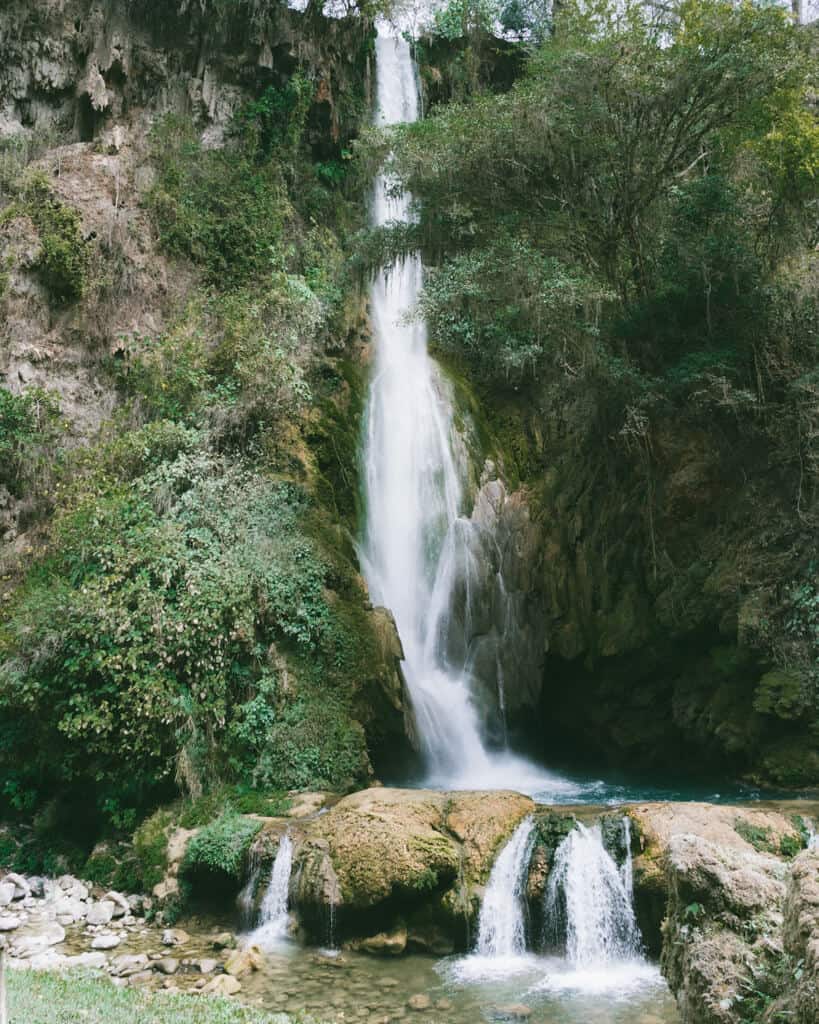 How to Visit Cascada El Aguacate: San Luis Potosi, Mexico 13 A tall, multi-tiered waterfall cascades down mossy rock formations into a shallow pool at Cascada El Aguacate in Huasteca Potosina, surrounded by lush greenery and rugged limestone cliffs.