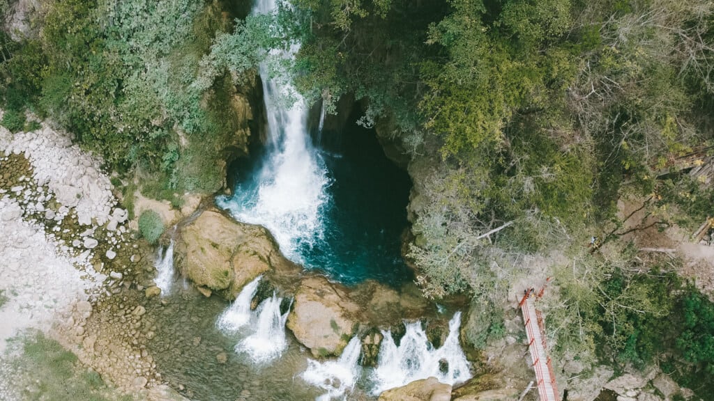 How to Visit Cascada El Aguacate: San Luis Potosi, Mexico 2 Aerial view of Cascada El Aguacate in Huasteca Potosina shows water plunging into a deep turquoise pool surrounded by lush forest. Smaller cascades flow downstream beside a red footbridge and rocky riverbank.