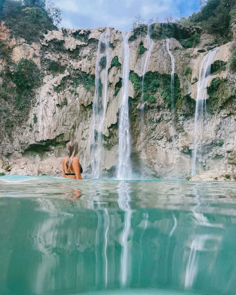 A shot of Cascada el Salto in Huasteca Potosina. The photo is partially underwater in the forground. With Sally standing in the water looking towards the waterfall dropping down a rocky canyon wall. 