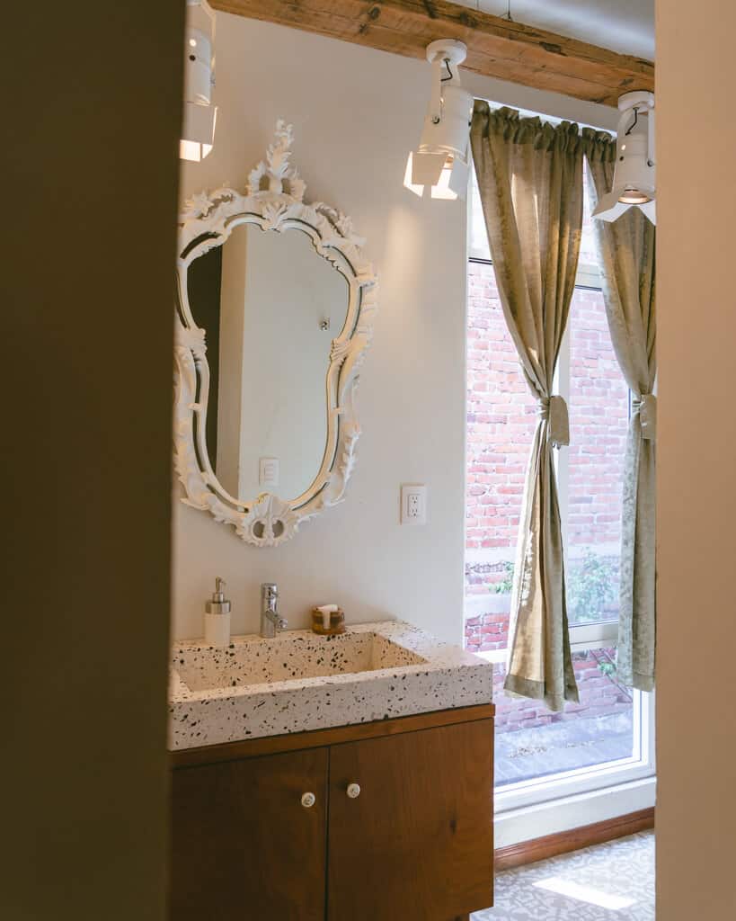 El Patio 77: Sustainable Bed and Breakfast Mexico City 7 A hotel bathroom with an ornate oval shaped mirror, terrazzo basin and floor tiles and green curtains tied open on a light window