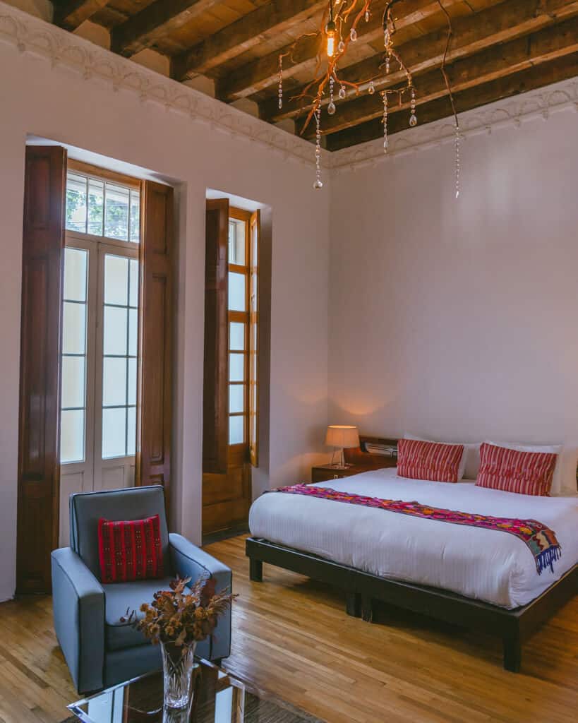 El Patio 77: Sustainable Bed and Breakfast Mexico City 9 A spacious historic style hotel room with a large bed with maroon cushions and blanket, a lounge chair and table with a vase of dried flowers at El Patio 77