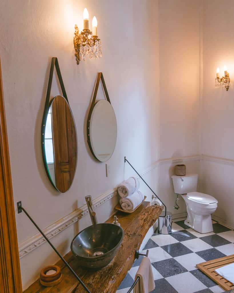 El Patio 77: Sustainable Bed and Breakfast Mexico City 10 A hotel bathroom with two round mirrors, a wooden vanity and black and white checked tiled floors with a toilet in the background