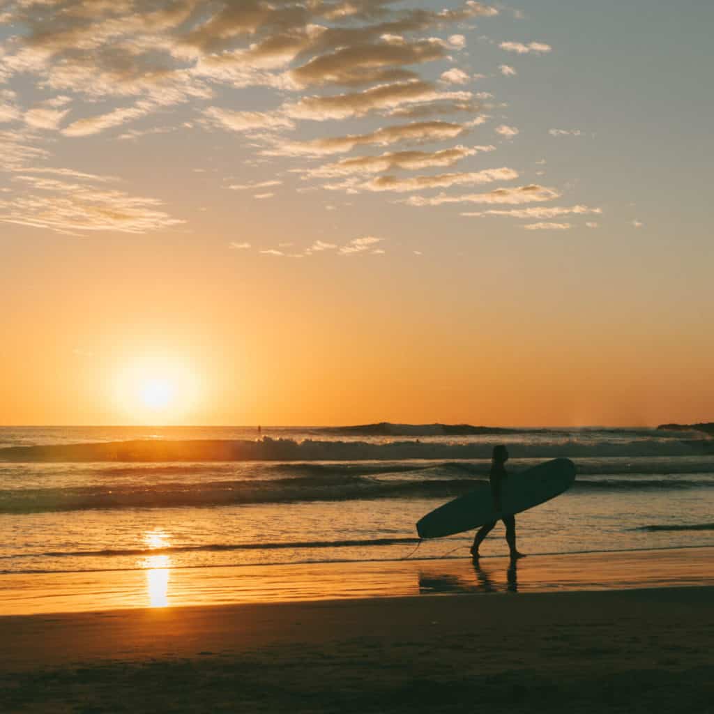 Cerritos Beach & El Pescadero, Mexico Travel Guide 7 Brayden carrying a board walks along the beach at sunset in Todos Santos, Mexico, with golden skies and gentle waves creating a tranquil scene.