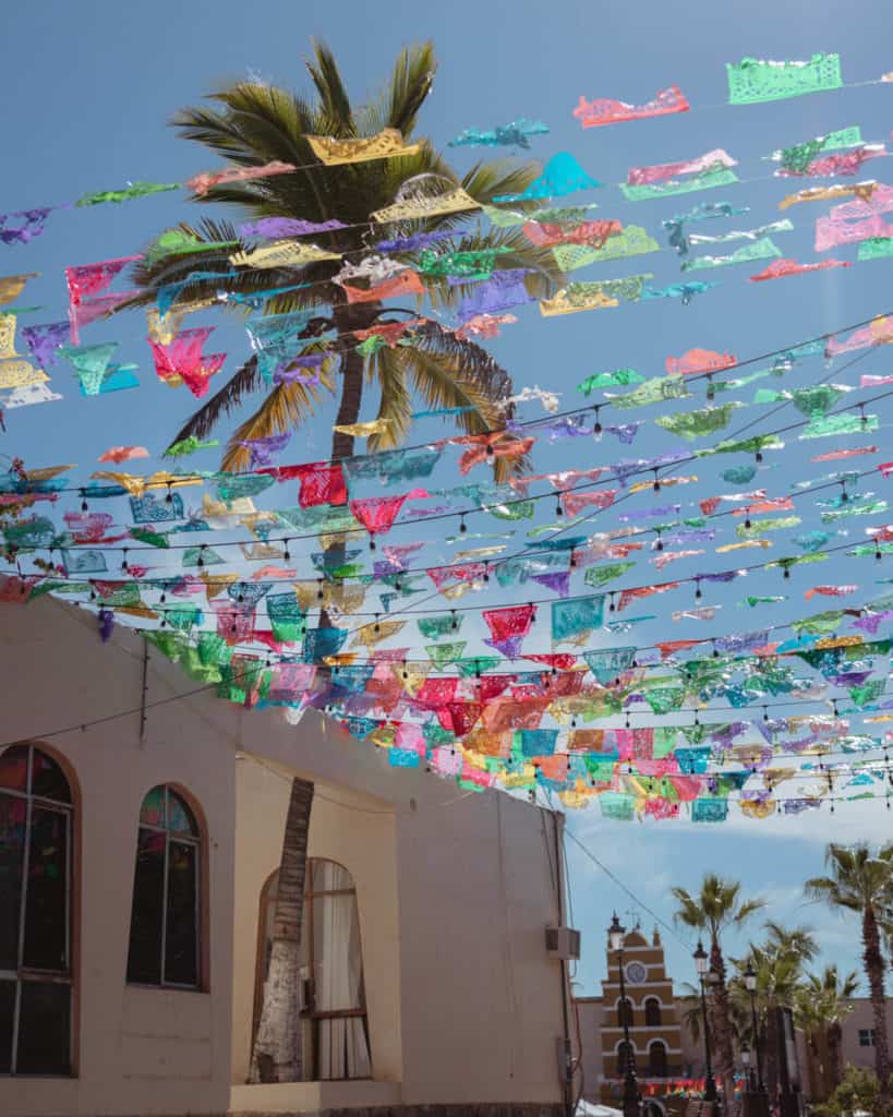 Where to Stay in Todos Santos, Mexico: Hotel & Area Guide 3 Colorful papel picado flags hang above a sunlit street lined with palm trees and colonial buildings, with the iconic Todos Santos church visible in the background in Todos Santos, Mexico.
