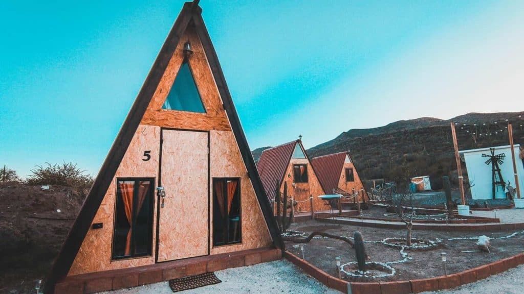 Mulege Mexico Travel Guide: Baja Sur's Oasis Town 19 A row of modern A-frame cabins in Indie Park with wood paneling and red roofs sits in a desert garden with cacti and string lights, backed by hills at sunset in Mulegé, Baja California Sur.
