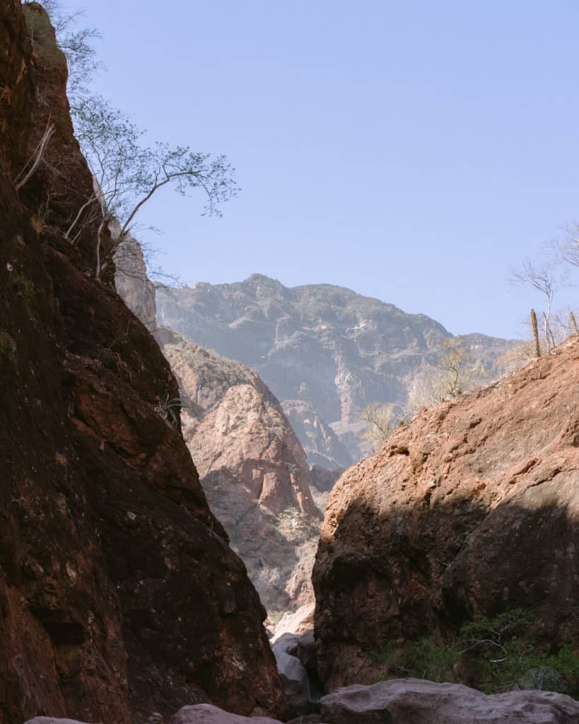 Tabor Canyon, Loreto Hiking Guide 7 A view from within a narrow canyon framed by steep red rock walls, opening up to reveal distant rugged mountains under a clear blue sky.