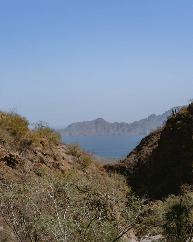 Tabor Canyon, Loreto Hiking Guide 12 A desert canyon in Loreto, Baja California Sur, opens up to a distant view of the Sea of Cortez and hazy mountain silhouettes under a clear blue sky.