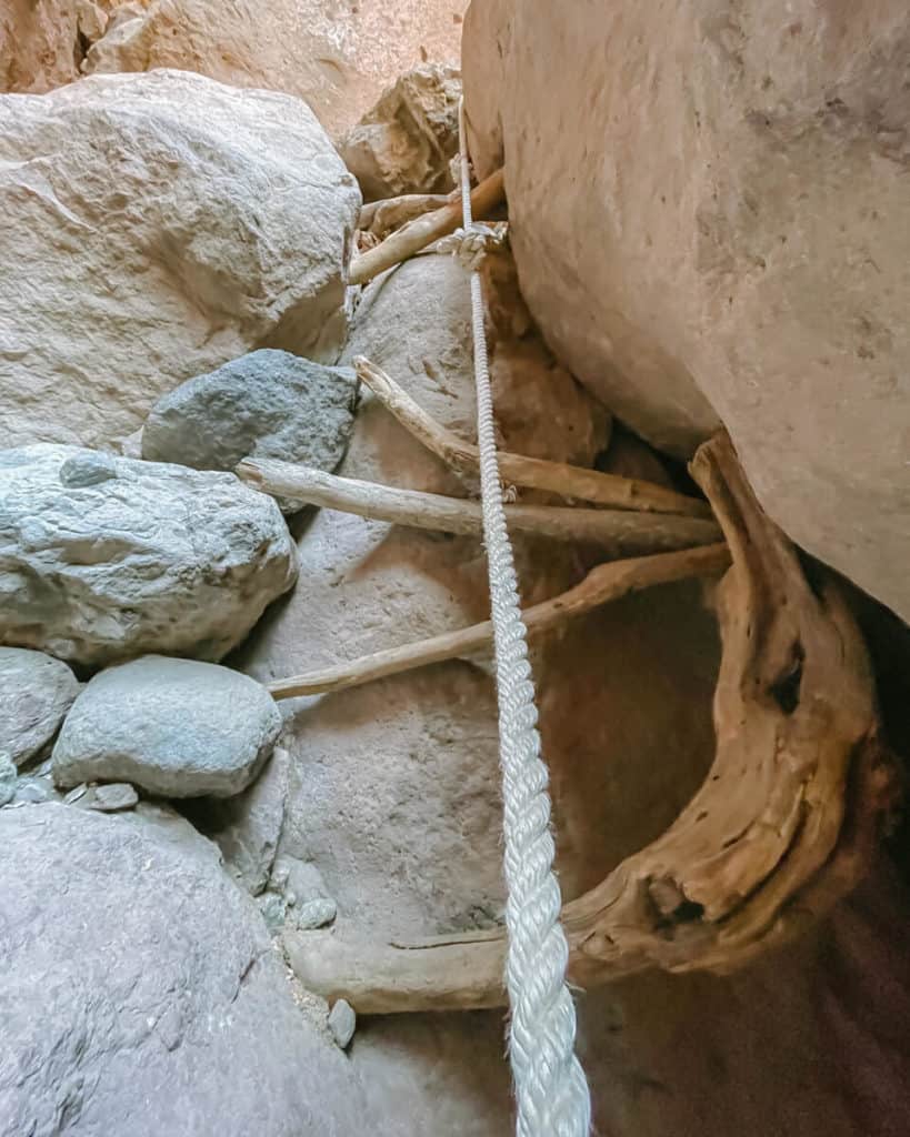 Tabor Canyon, Loreto Hiking Guide 11 A primitive ladder made of wooden branches and a yellow climbing rope rises between towering canyon boulders, framed by a clear blue sky above.