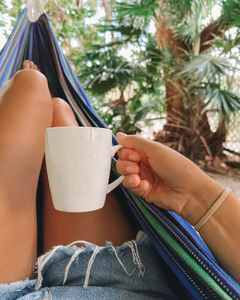 Our Belize Trip Cost: Is Belize Expensive? 30 Sally relaxes in a striped hammock in San Ignacio, Belize, holding a white ceramic mug, with her legs outstretched and surrounded by lush tropical greenery in the background.