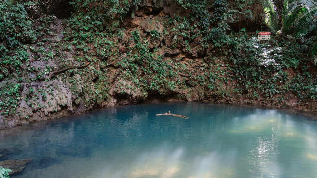 10 Top Things to do in Belize: Your Belize Bucket List 14 floats on a log in the center of the turquoise Blue Hole in San Ignacio, Belize, surrounded by lush greenery and steep rock walls, with a sign reading “Danger. Don’t Jump.” in the background.