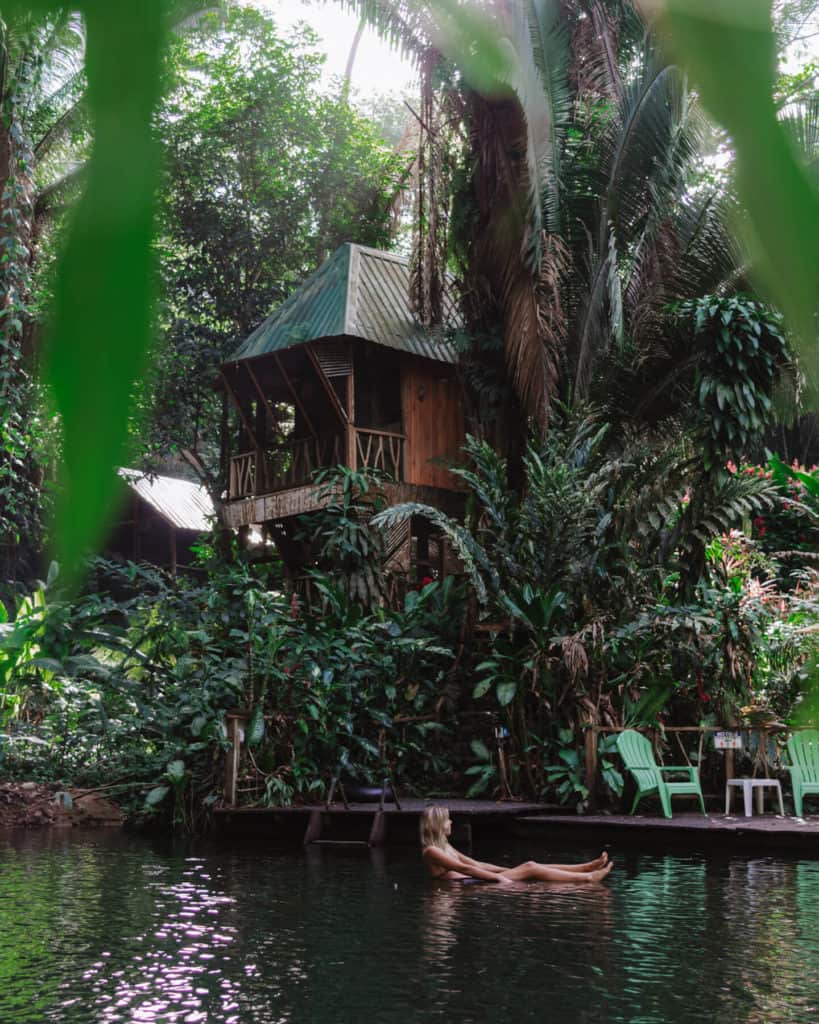 10 Top Things to do in Belize: Your Belize Bucket List 17 Sally floats peacefully in a jungle river in San Ignacio, Belize, near a rustic wooden treehouse surrounded by lush tropical plants and tall palm trees, with green Adirondack chairs on the riverbank.