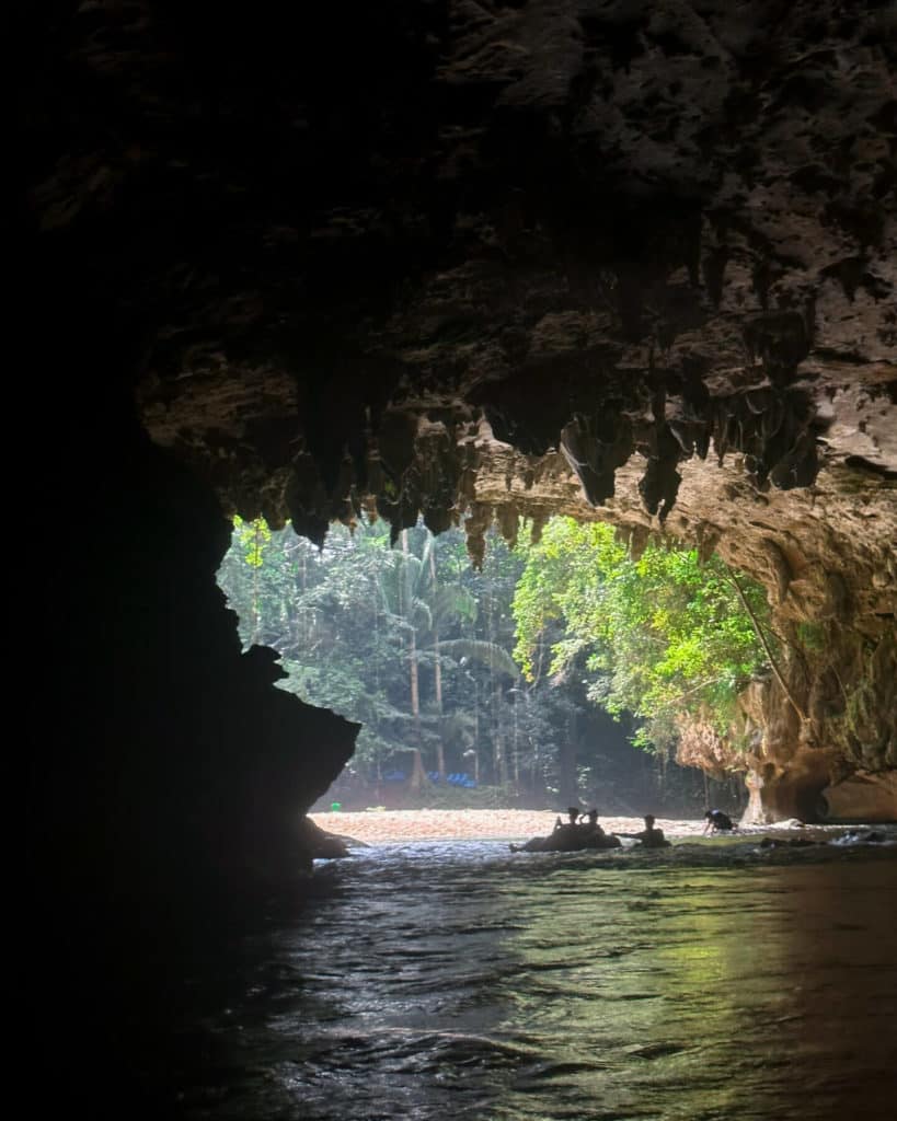 10 Top Things to do in Belize: Your Belize Bucket List 9 Silhouetted figures of three people, possibly including Sally and Brayden, stand at the mouth of a large cave near San Ignacio, Belize, with dense green jungle trees visible beyond the cave’s opening.