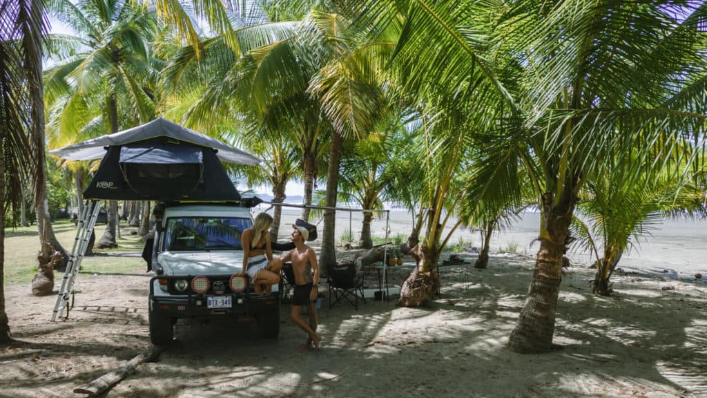 Sally and Brayden leaning on a white 4x4 with a rooftop tent set up amongst palm trees beach camping in Costa Rica