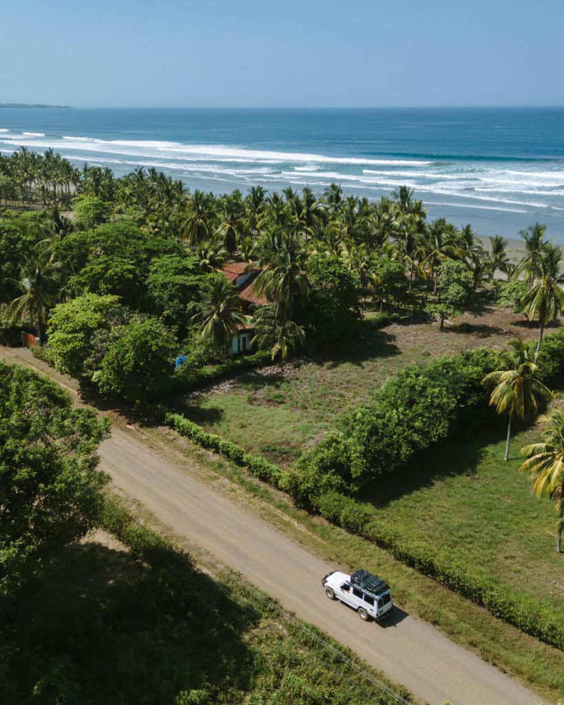 An aerial shot of a white land cruiser driving down a dirt road near the beach lined with palm trees in Costa Rica
