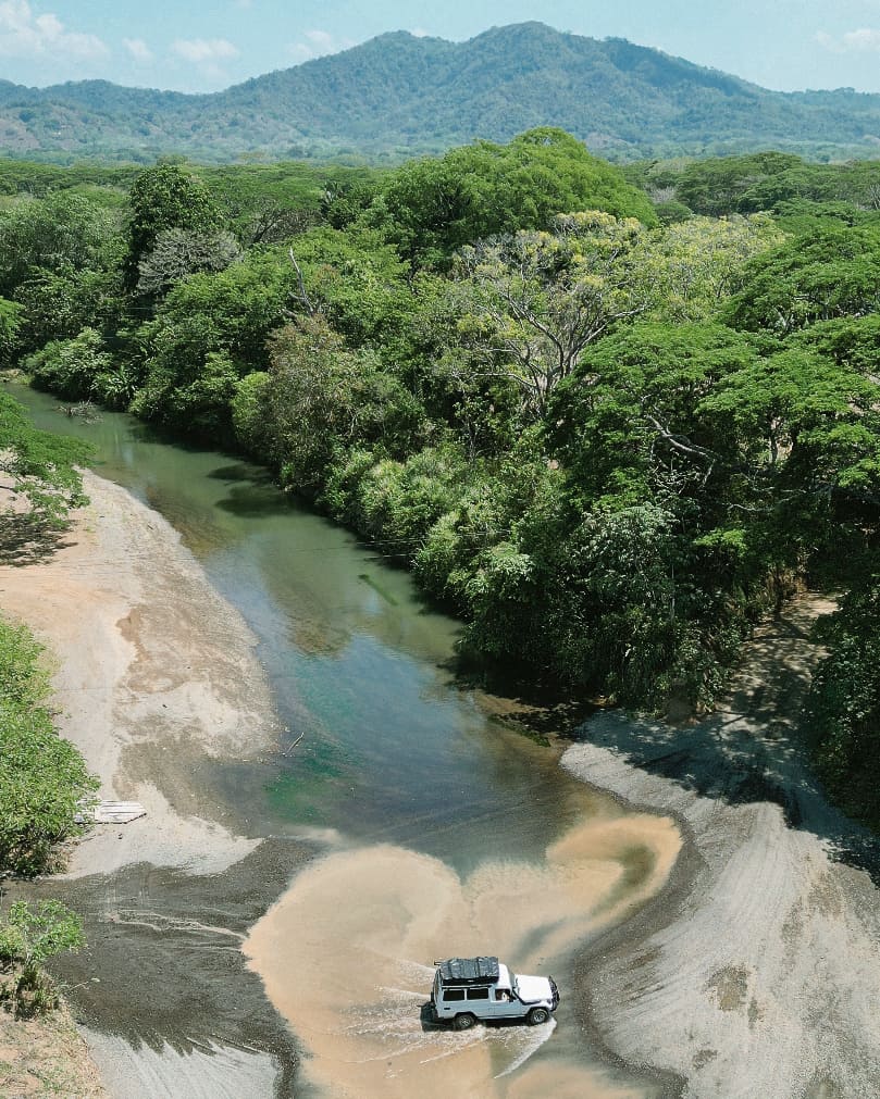 A drone shot of a white 4x4 driving across a river in Costa Rica with mountains in the background