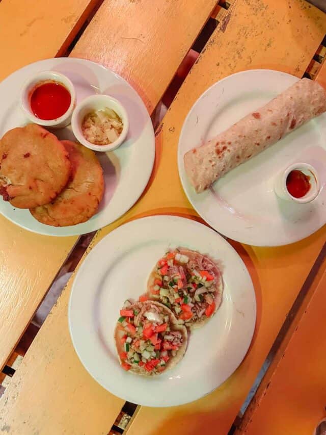 Our Belize Trip Cost: Is Belize Expensive? 18 Three plates of traditional Belizean food on a yellow wooden table, including stuffed fry jacks with hot sauce and pickled onions, a flour tortilla with sauce, and mini garnaches topped with chopped