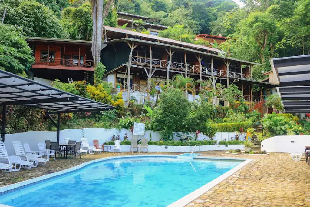 19 Best Things to Do in Manuel Antonio, Costa Rica 33 Multi-level wooden hotel built into a lush hillside with balconies overlooking a stone patio and rectangular outdoor pool, surrounded by white lounge chairs, palm trees, and tropical landscaping.