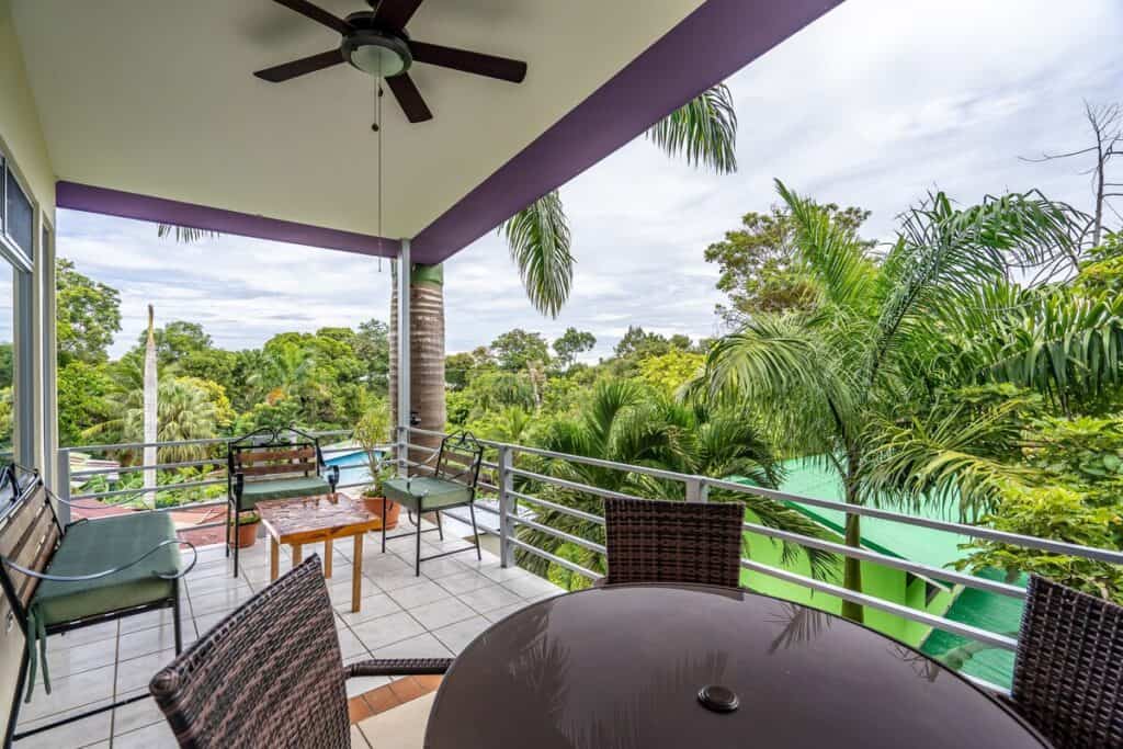 19 Best Things to Do in Manuel Antonio, Costa Rica 34 Peace Paradise Hotel's covered balcony with ceiling fan and patio furniture overlooking a dense tropical landscape filled with palm trees and a distant glimpse of a pool. The seating area includes wooden benches with green cushions and a round table with wicker chairs on a tiled floor.