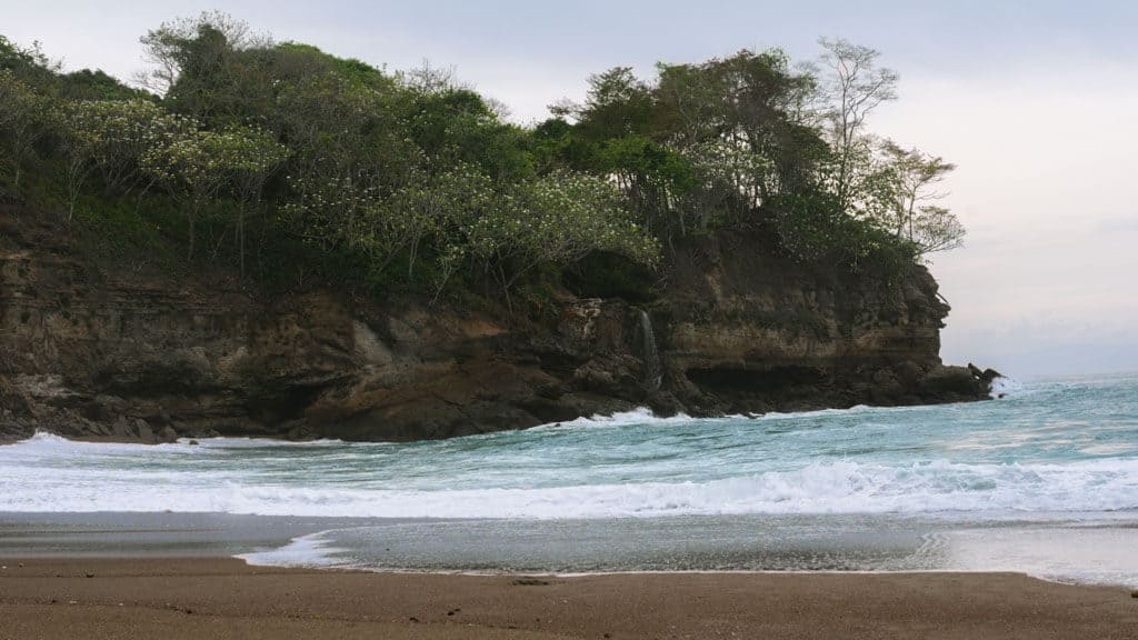 A wide shot of El Chorro Waterfall in the distance from Playa Cocalito, with the sand and shoreline in the foreground and the waterfall and cliff in the background