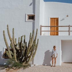 Sally standing in front a white washed building with a large cactus plant growing next to her with multiple cacti heads branching out in all directions