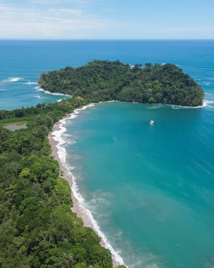 An aerial shot of Punta Catedral, a jungle clad rocky point in Manuel Antonio, connected to the mainland via a narrow land bridge, with two bays and beaches on either side 