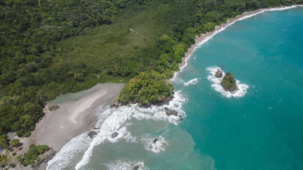 A drone shot of the coastline in Manuel Antonio, where green forest meets bright blue ocean, with small rocky outcrops dotting the water