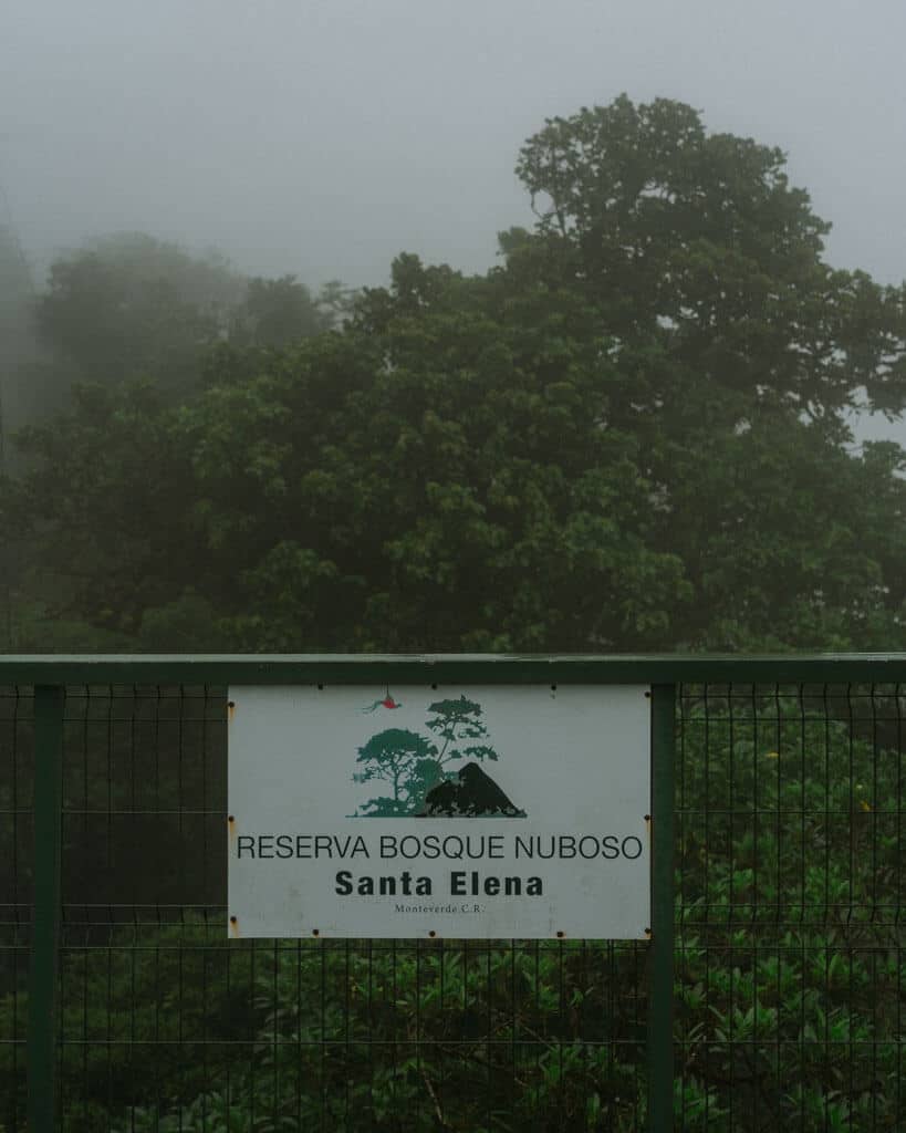 A sign on a green metal lookout platform with the worse 'Reserva Bosque Nuboso Santa Elena' with fog and trees in the background