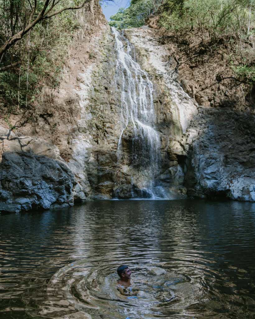 15 Best Things to Do in Montezuma Costa Rica 29 Brayden floating in the pool below the Montezuma Waterfall, with a small cascade of water dropping down a jagged rocky cliff behind him
