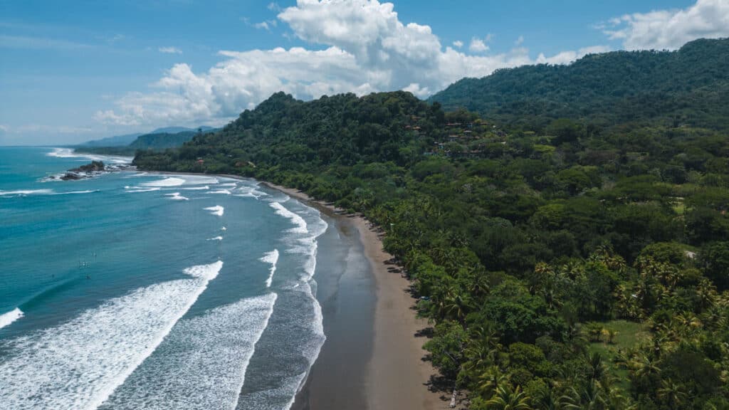 Aerial view of Dominical's rugged coastline with surfers in the water, the beach curving between the lush tropical greenery and the Pacific Ocean.
