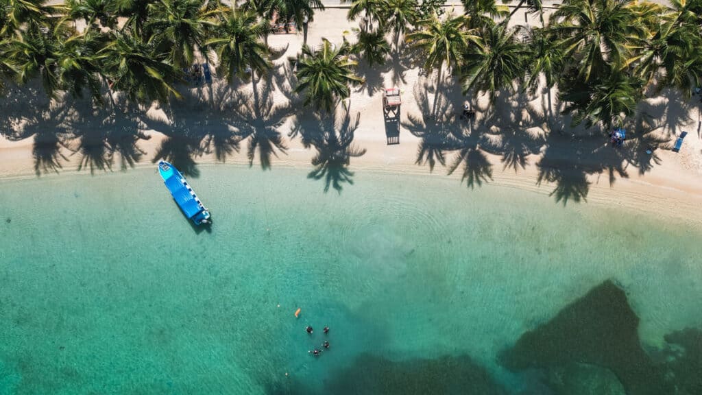 18 Things to Do in Roatan (+ Unethical Things NOT to Do) 18 Overhead shot capturing the tropical landscape of Half Moon Bay, Roatan, with a blue boat resting by the palm-lined beach.