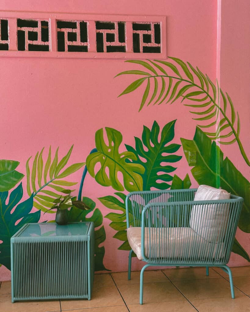 Our Top 10 Restaurants in Utila, Honduras 8 A stylish corner of Josie's Cafe in Utila with tropical vibes, showcasing a pink wall with leafy plant murals and chic, blue-green modern furniture.