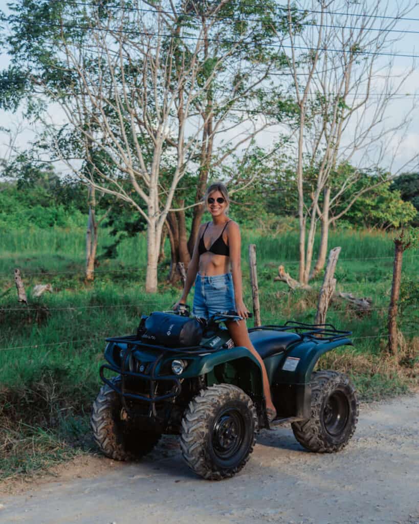 Sally in a black bikini top and shorts posing on an ATV on a dirt path with tropical vegetation and trees in the background, symbolizing adventure and exploration of Utila
