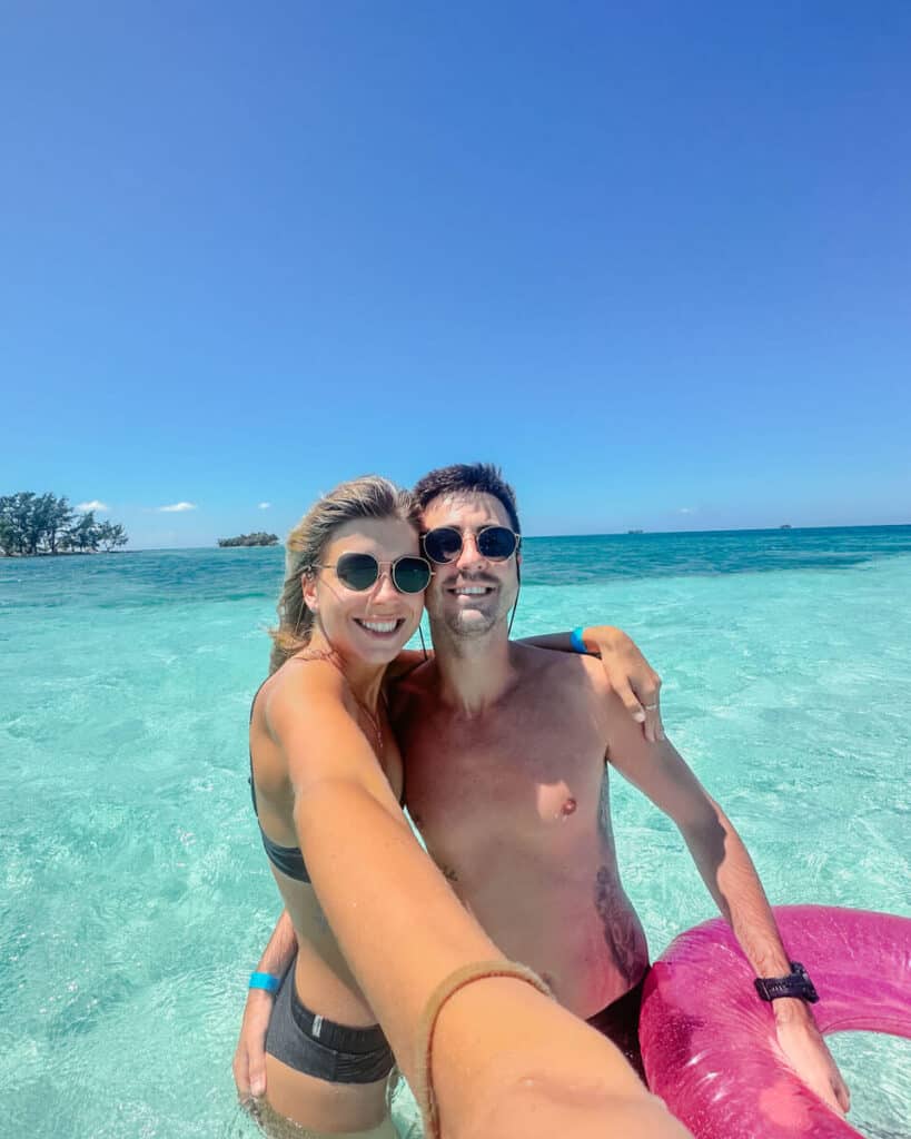 How to Visit Water Cay Utila: Untouched Tropical Heaven 20 Sally and Brayden take a joyful selfie in the crystal-clear waters of Water Caye, with vibrant blue skies and lush greenery in the background, capturing a moment of tropical bliss