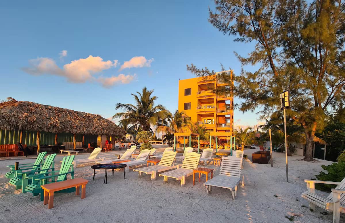 Where to Stay in Caye Caulker: 16 Options For All Budgets 19 A vibrant beachside scene at Bay Towers Hotel in Caye Caulker, Belize, featuring a bold orange building, rows of white lounge chairs and colorful Adirondack chairs on the sand, with palm trees and a thatched-roof cabana creating a relaxed tropical vibe at sunset