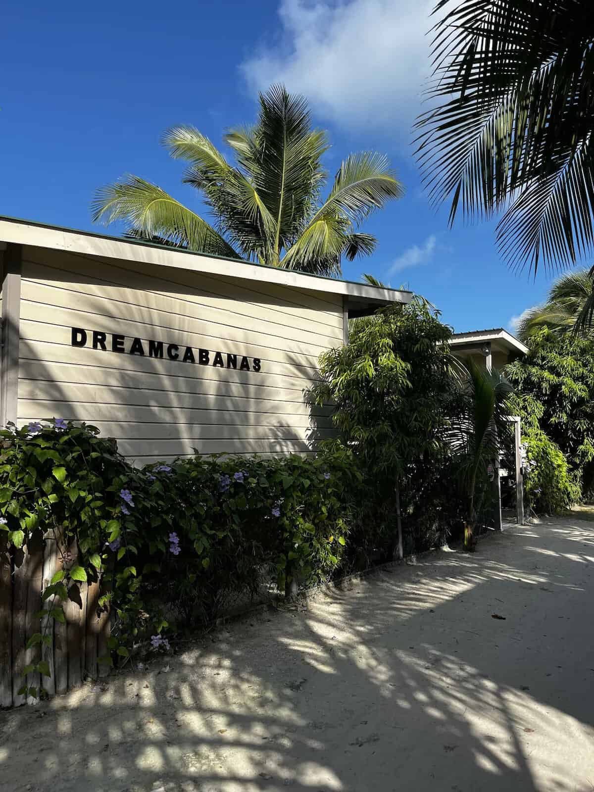 Where to Stay in Caye Caulker: 16 Options For All Budgets 12 A light-colored building with the sign "DREAMCABANAS" in Caye Caulker is surrounded by lush tropical plants and palm trees under a bright blue sky. The sandy path and shadows from the foliage enhance the tranquil, island atmosphere.