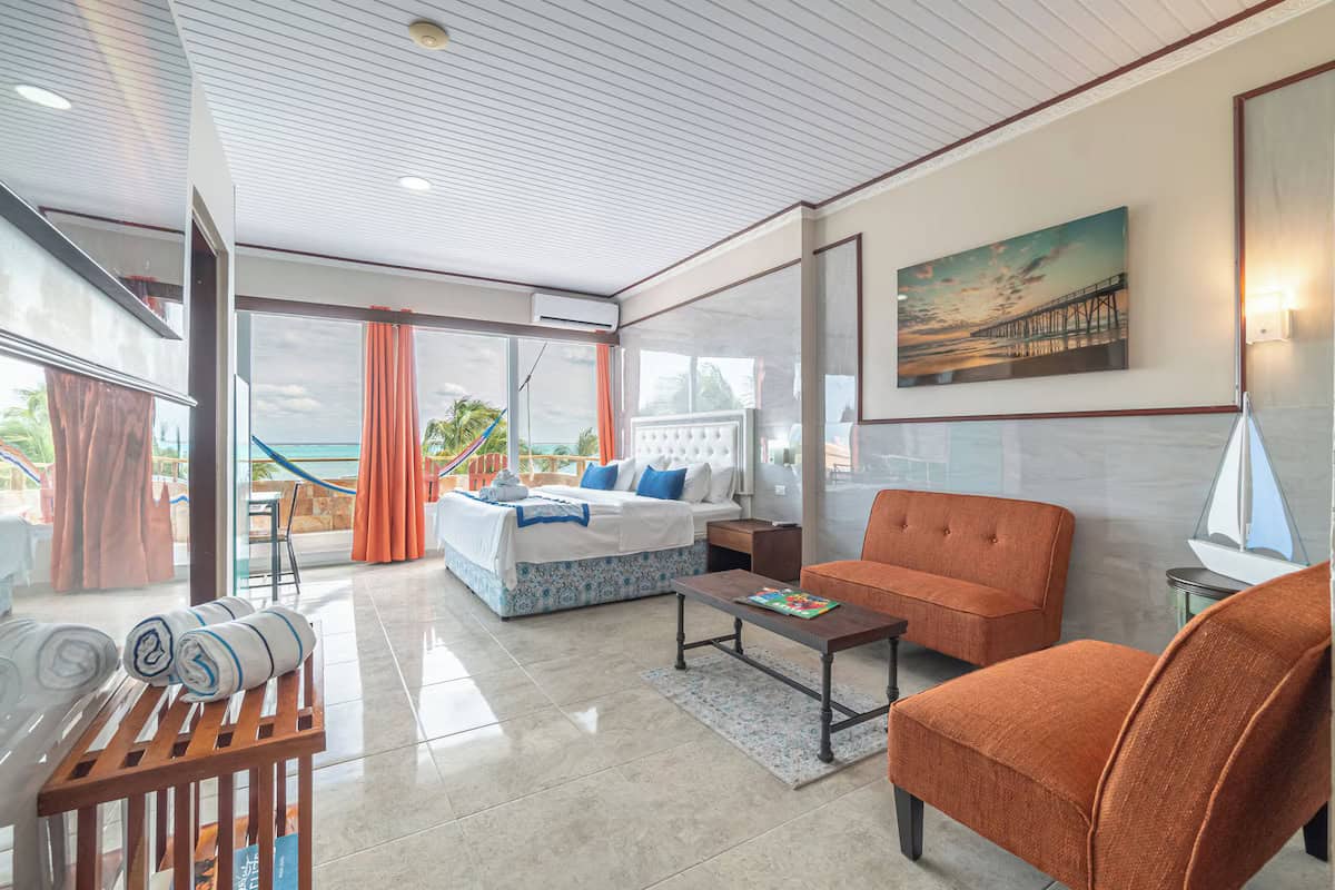 Where to Stay in Caye Caulker: 16 Options For All Budgets 15 A bright and airy hotel room in Island Magic Beach Resort in Caye Caulker with modern decor, featuring a king-size bed, orange lounge chairs, and large windows opening to a balcony with ocean views and hammocks.