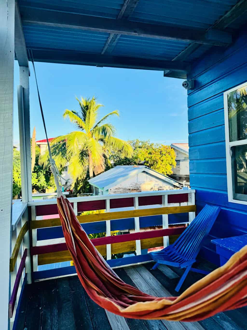 Where to Stay in Caye Caulker: 16 Options For All Budgets 9 A cozy balcony in Go Slow Hostel Caye Caulker features a striped hammock and a blue wooden lounge chair, with a colorful railing and a bright blue wooden house wall. Palm trees and rooftops are visible under a clear blue sky.