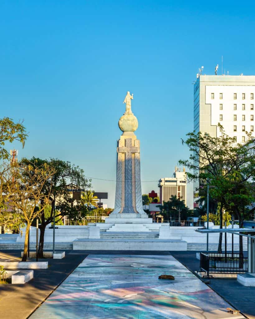 Complete El Salvador Travel Guide 16 The Monument to the Divine Savior of the World (Monumento al Divino Salvador del Mundo) in San Salvador, El Salvador, featuring a statue of Jesus Christ atop a globe and tall pedestal, with surrounding city buildings and a colorful mural on the ground in the foreground.