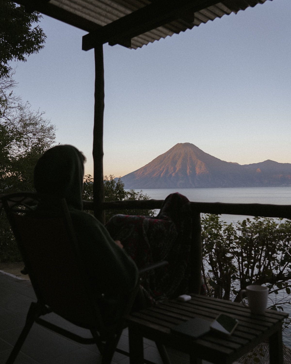 Lake Atitlan Travel Guide: Everything You Need to Know 12 Brayden sitting in a chair on a covered patio, enjoying a breathtaking view of a volcano and Lake Atitlan. The scene captures the serene morning atmosphere with the sun casting a soft light on the landscape.