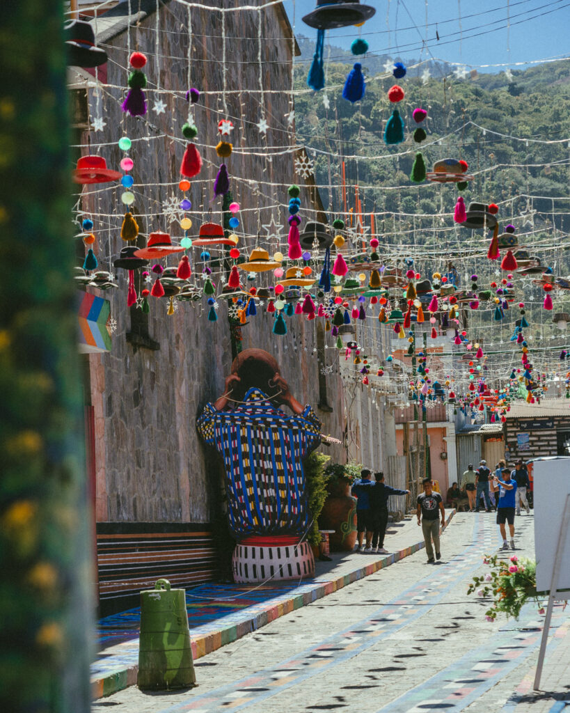 Lake Atitlan Itinerary: How to Spend 7, 5 or 3 Nights 22 A narrow street in San Juan adorned with hanging colorful decorations. A large mural of a traditional Maya person with a basket on their head is seen on one of the walls.