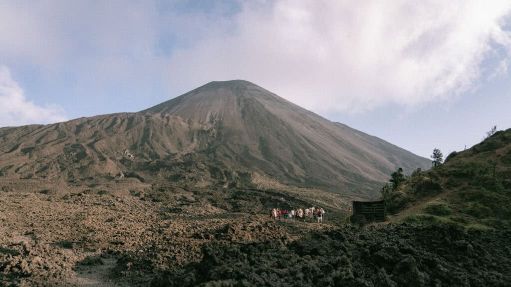 A wide landscape showing a group of tourists on a guided tour at the base of Volcan Pacaya. The volcano looms large in the background with a barren, rocky landscape stretching out in front.