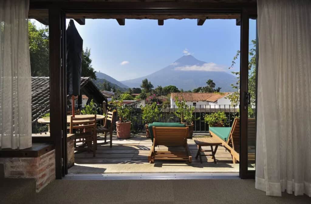Where to Stay in Antigua Guatemala: Best Boutique Stays 28 A wooden rooftop deck at Casa Santa Domingo in Antigua Guatemala, with lounge chairs and a dining set with umbrella overlooking Volcan Agua, its summit covered in cloud.