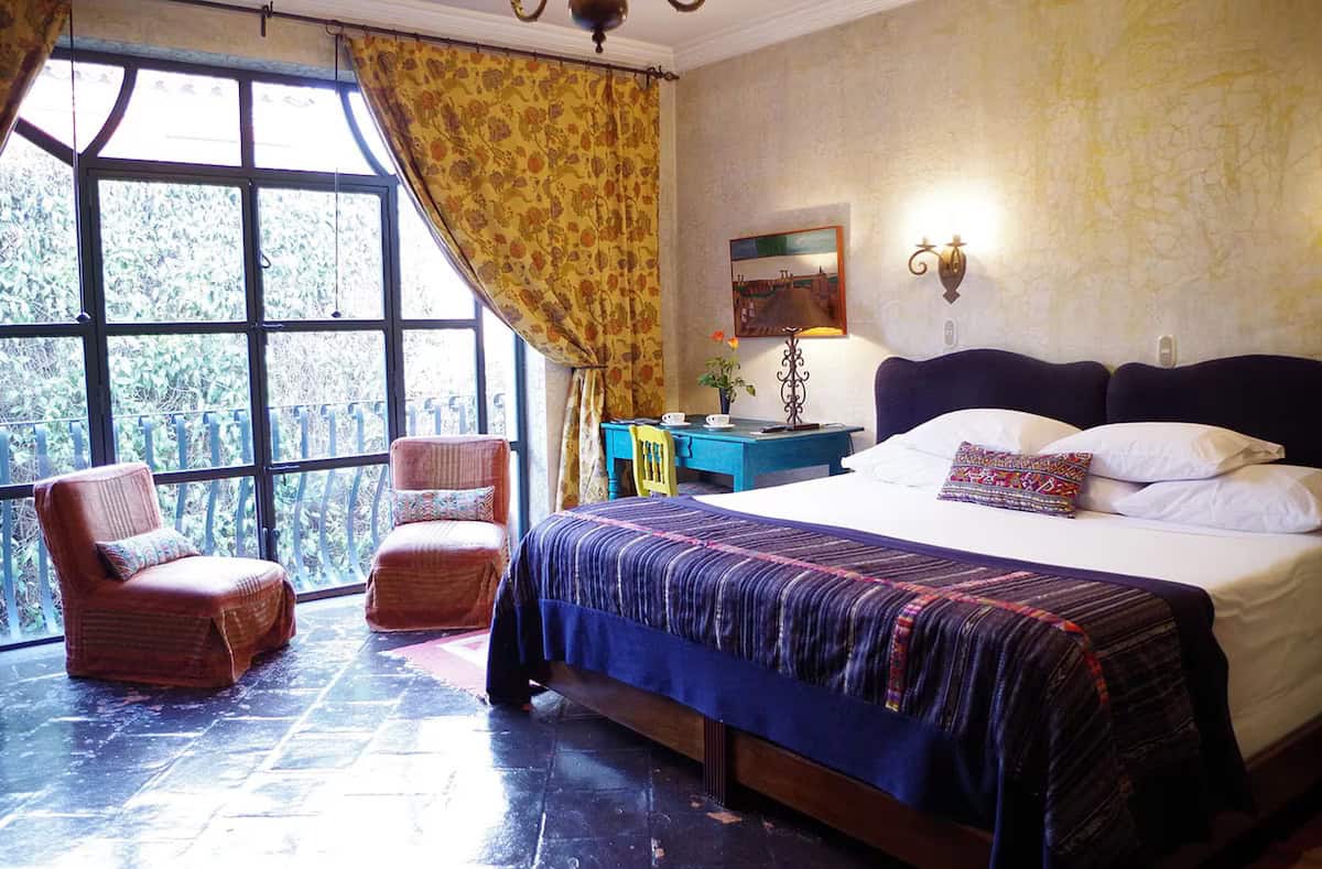 Where to Stay in Antigua Guatemala: Best Boutique Stays 30 A grand and sophisticated bedroom at Meson Panza Verde, where to stay in Antigua Guatemala. Featuring a lit fireplace, a grand chandelier, dark wooden furniture and big glass doors opening to a courtyard with a fountain and plants.