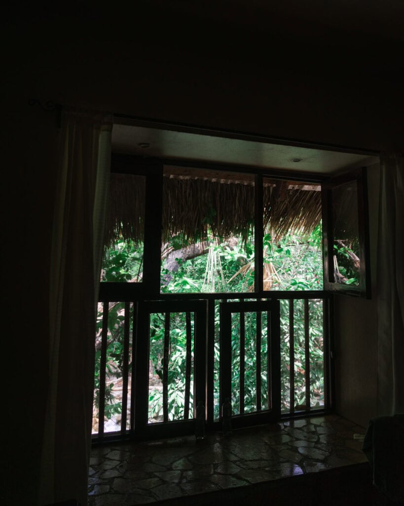 Cascadas El Chiflon: Without a Tour + Overnight Stay 10 View of a large window framed by curtains inside a cabin at Cascadas El Chiflon. Through the window, lush green trees and tropical foliage can be seen outside, blending into the jungle-like surroundings.