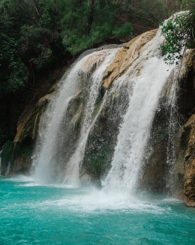 Cascadas El Chiflon: Without a Tour + Overnight Stay 17 A stunning view of Cascada Ala de Angel, a wide waterfall tumbling over rocky cliffs into a turquoise pool at the base, surrounded by green trees and vegetation.