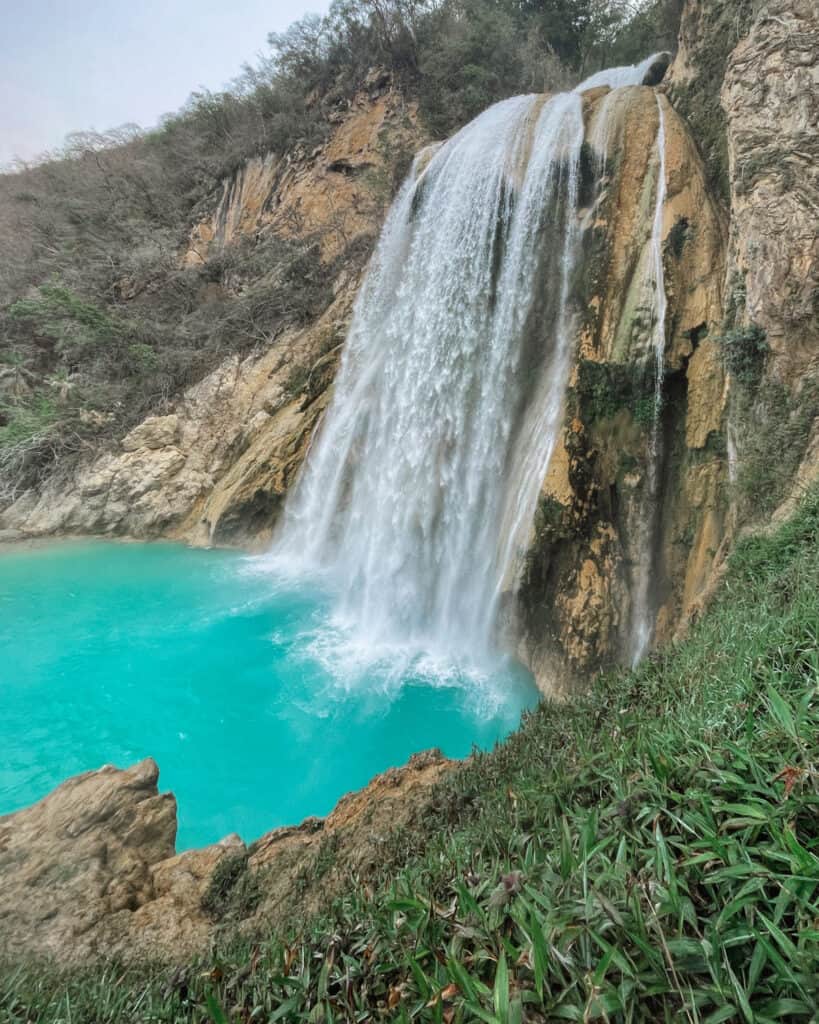 Cascadas El Chiflon: Without a Tour + Overnight Stay 24 A close-up of Cascada Arcoiris, a powerful waterfall crashing down rocky cliffs into a vivid turquoise pool. The surrounding greenery contrasts with the natural rock formations.