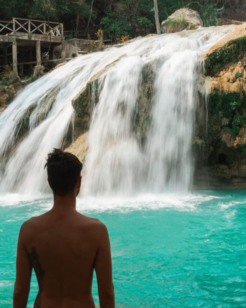 Cascadas El Chiflon: Without a Tour + Overnight Stay 15 Brayden is standing shirtless, facing the waterfall at Cascada El Suspiro. He observes the water crashing down into the turquoise pool, with a wooden platform visible in the background.